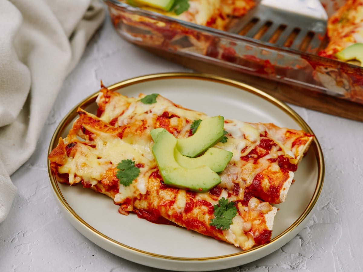 a side view of a plate with six baked enchilada rolls topped with cheese, cilantro, and avocado slices