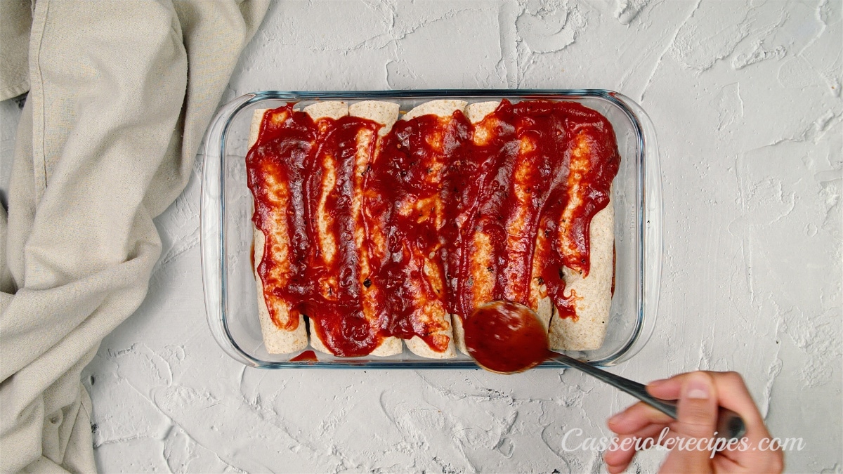 an overhead view of a glass baking dish with enchilada rolls and a hand spreading red sauce on top with a spoon