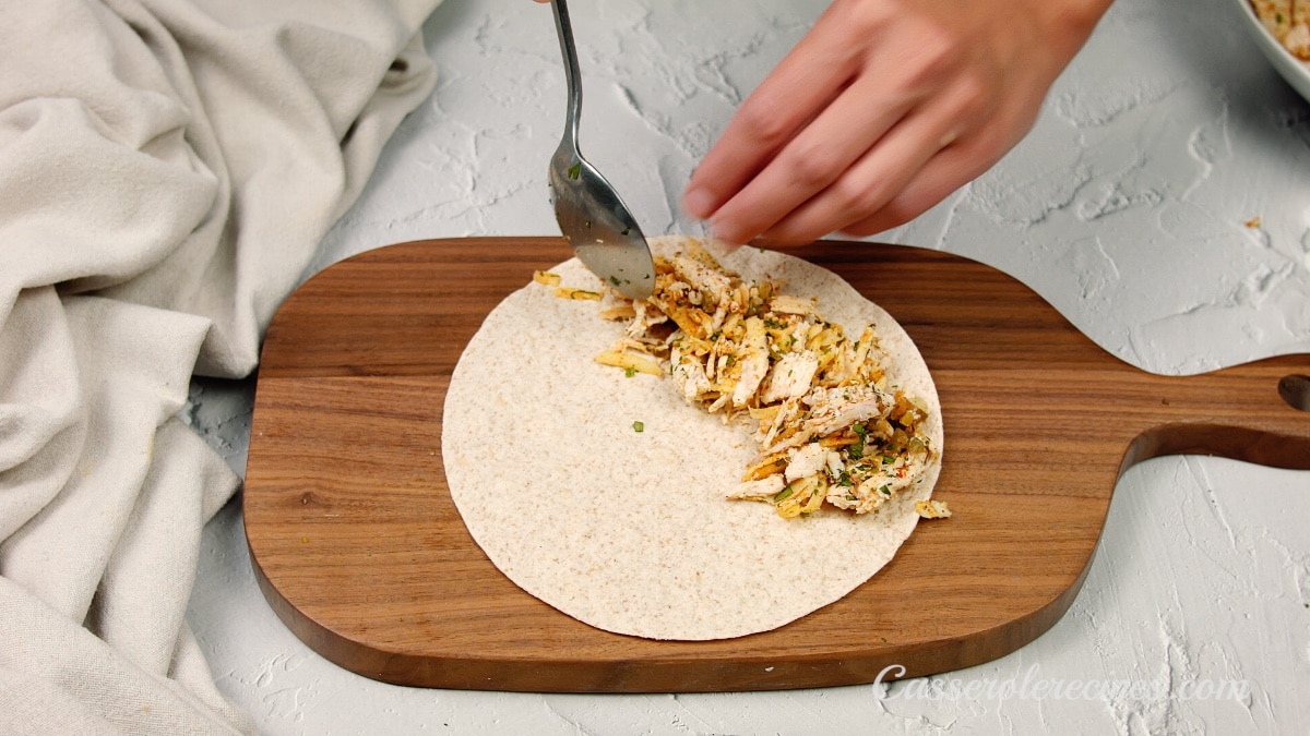 a flat tortilla being filled with chicken by a hand and a fork on a wooden cutting board