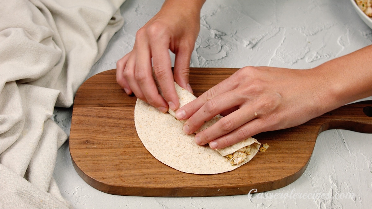 two hands rolling an enchilada on a wooden cutting board