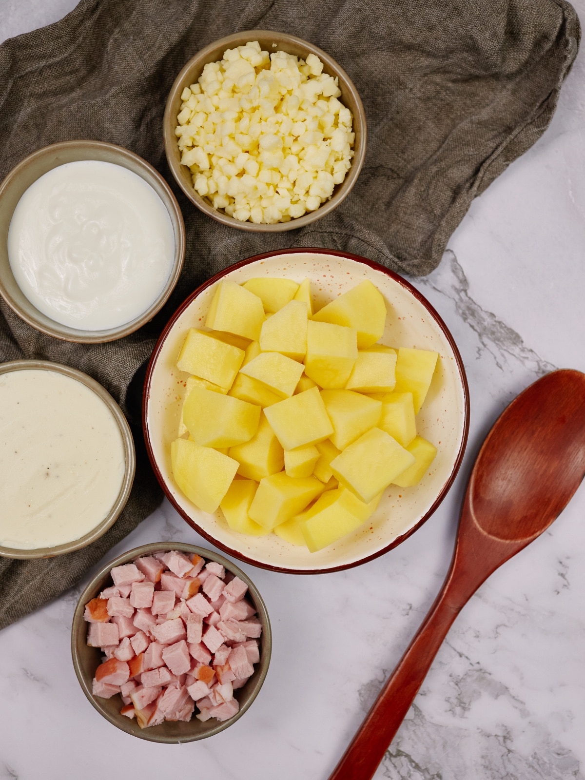 ingredients for ham and potato casserole in bowls next to a wooden spoon on a marble counter