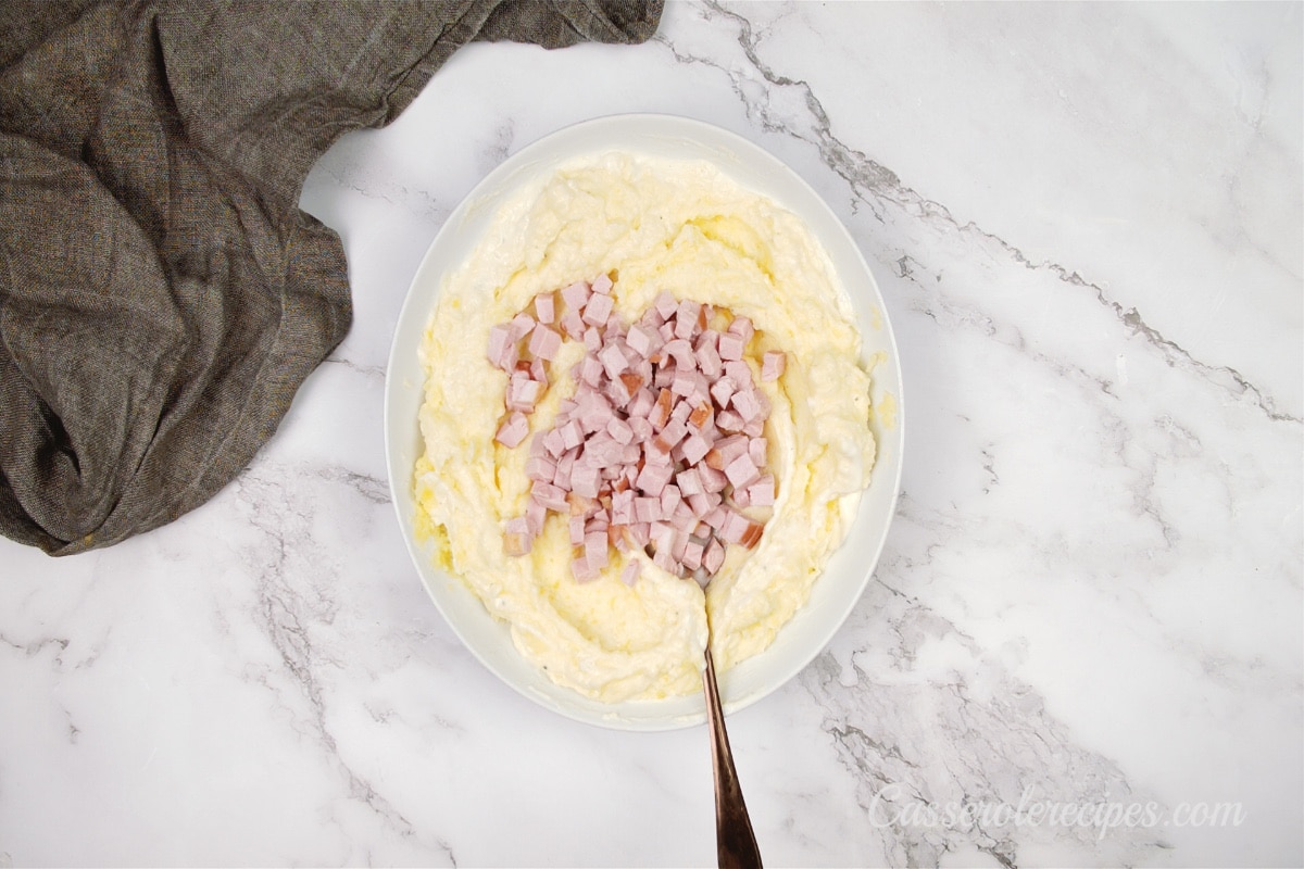 ham added to the top of a bowl of mashed potatoes in a white bowl on a marble surface