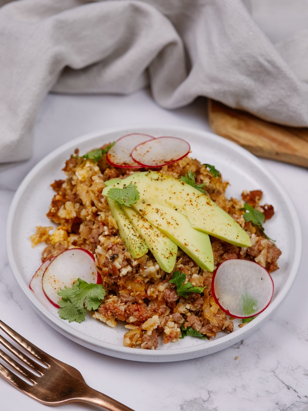 a side view of keto taco casserole on a white plate topped with sliced radishes, avocados, and cilantro leaves