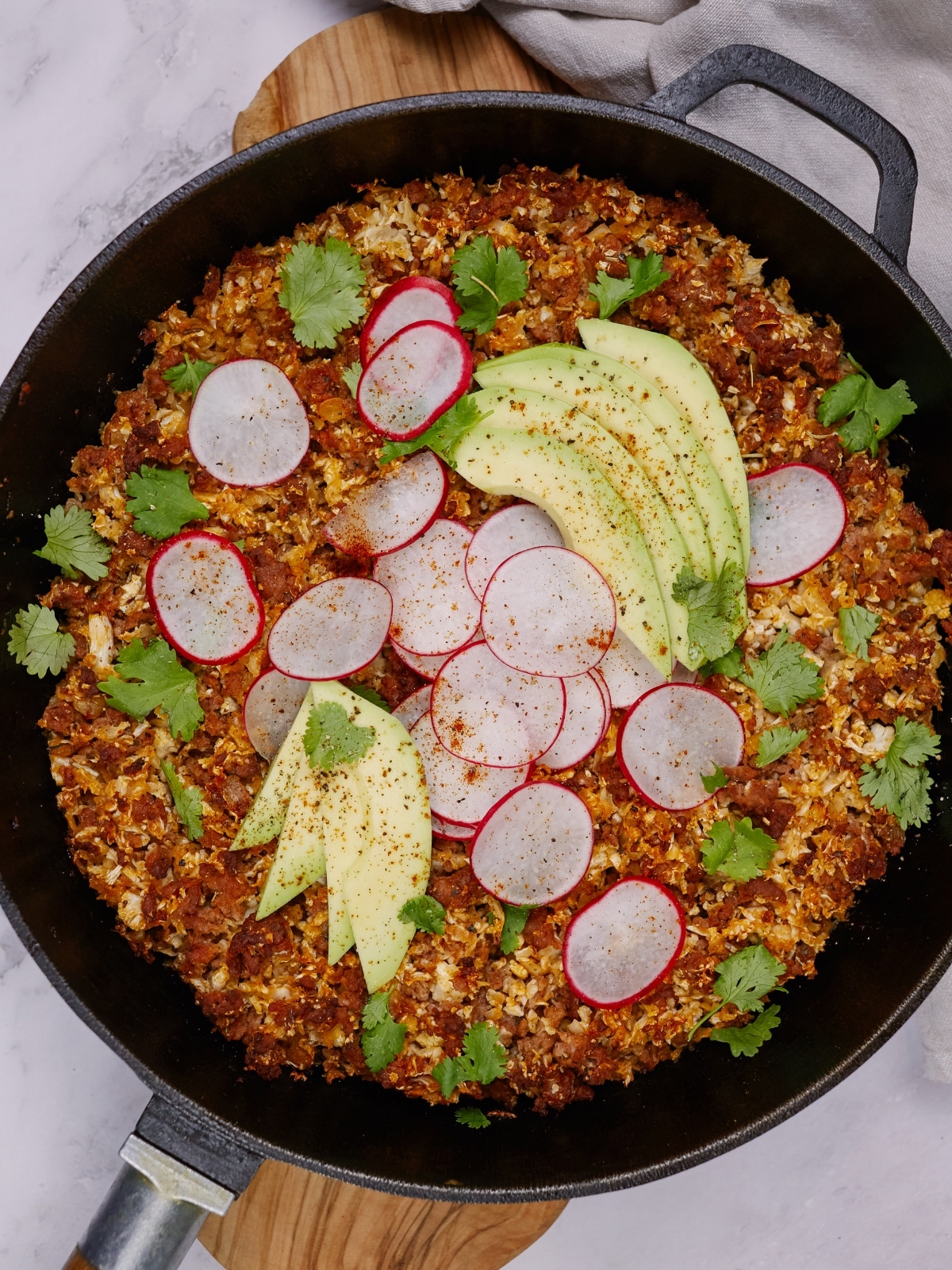a cast iron skillet with keto taco casserole topped with sliced radishes, avocados, and cilantro leaves