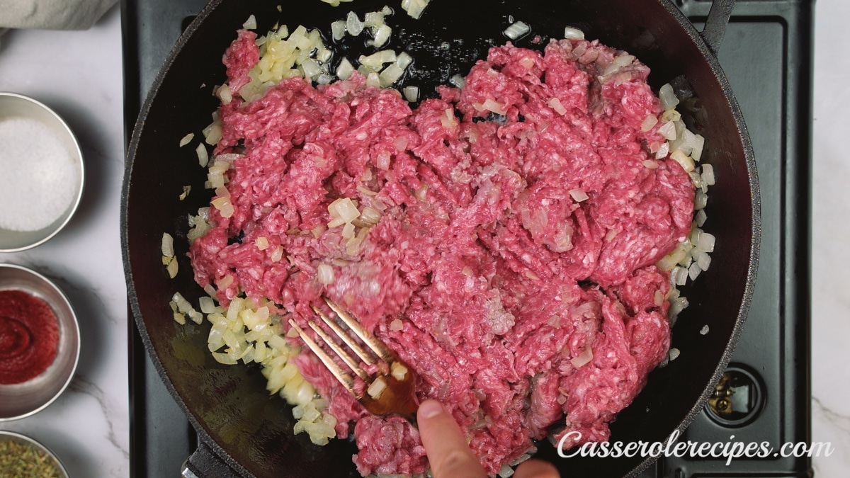a pan with cooked onions and raw ground beef being mixed in with a fork