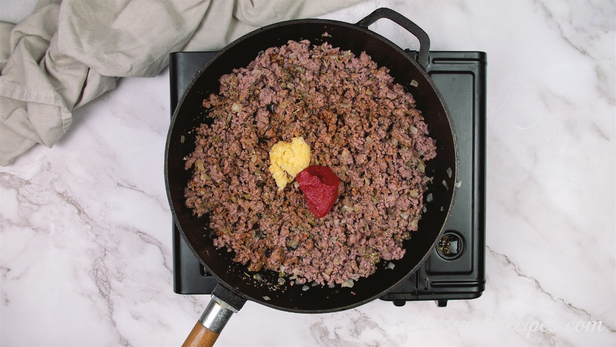 a pan of cooked ground beef on the stove topped with tomato paste and minced garlic