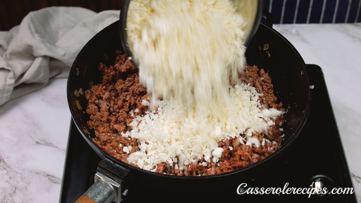cauliflower rice being poured into the cast iron skillet filled with ground beef and seasonings