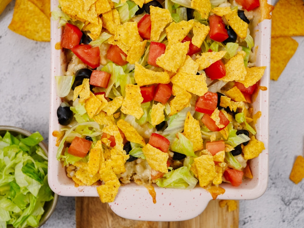 Mexican casserole in a white baking dish topped with tortilla chips, tomatoes, olives, and lettuce