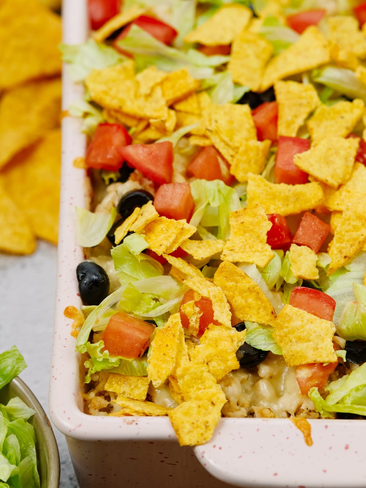 Mexican casserole in a white baking dish topped with tortilla chips, tomatoes, olives, and lettuce