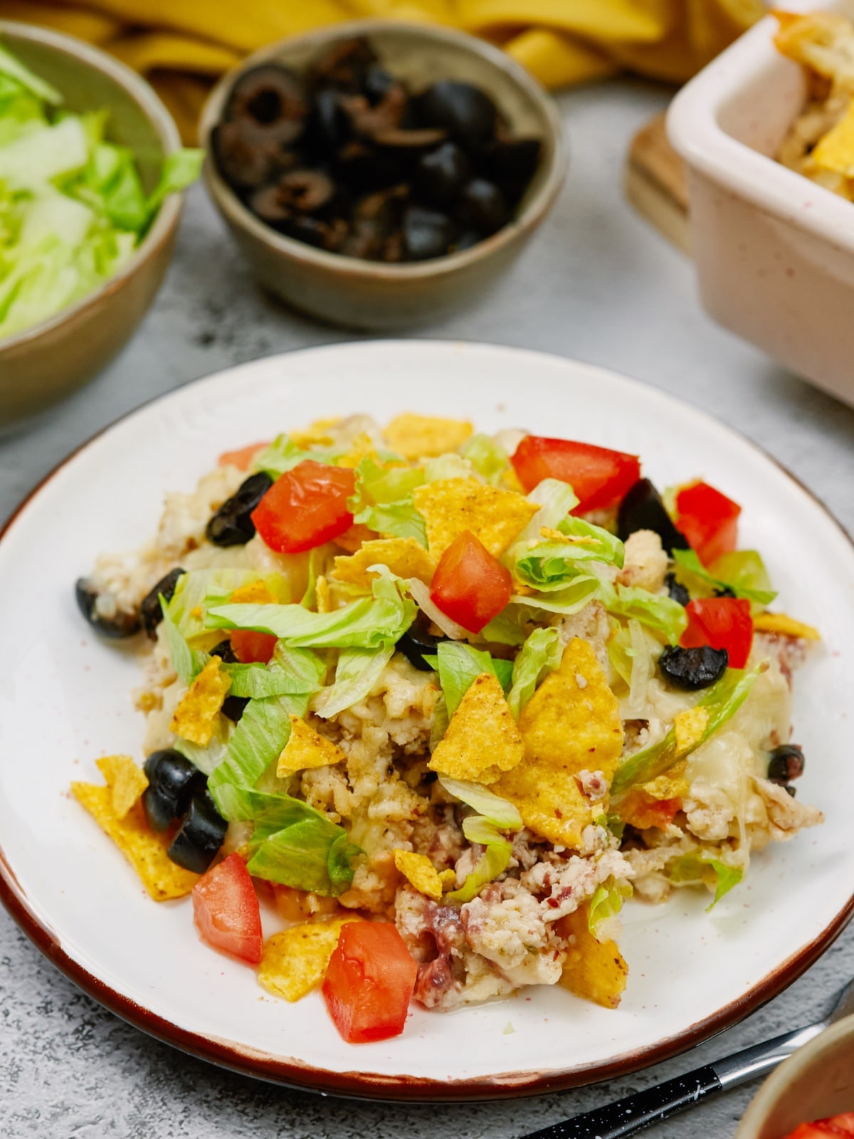 a plate of casserole topped with tortilla chips, olives, lettuce, and tomatoes