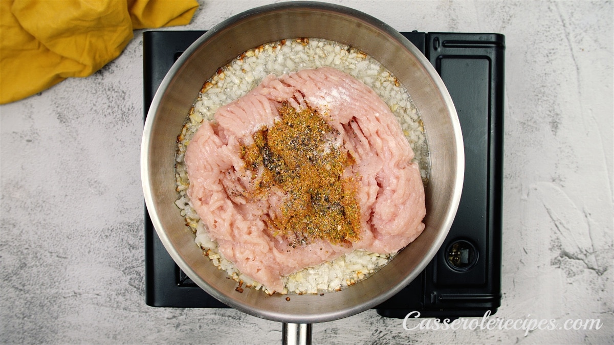onions and raw ground chicken topped with spices in a pan on a stove top