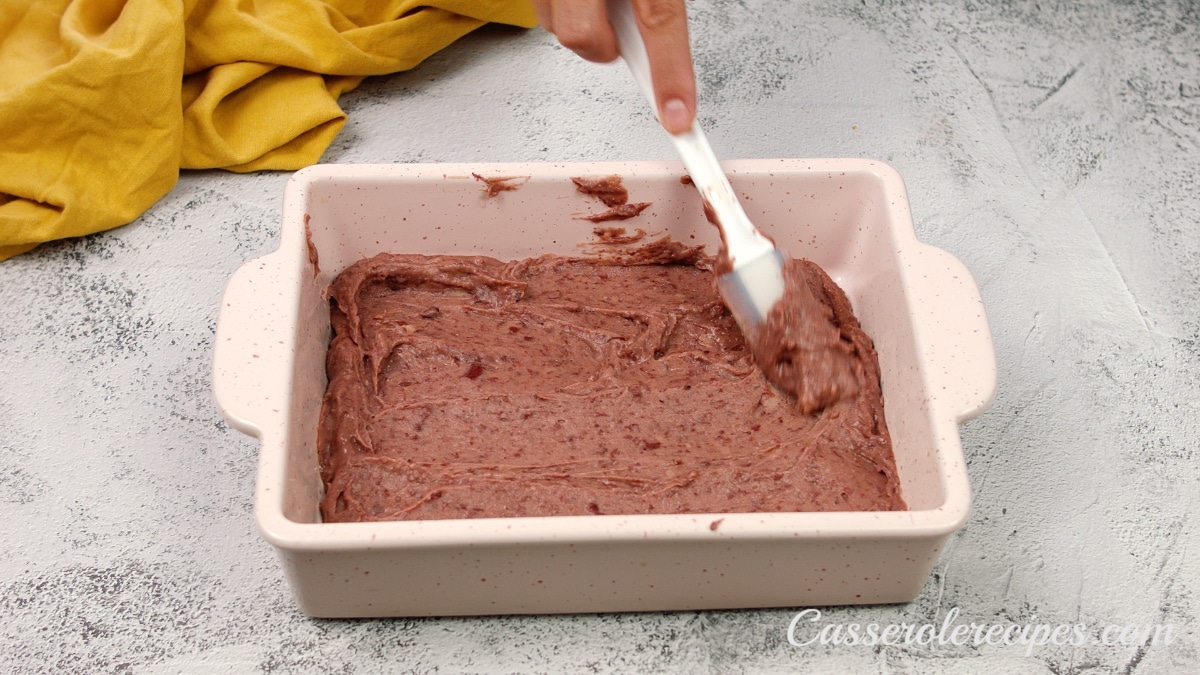 refried beans being spread on the bottom of a white casserole dish with a spatula