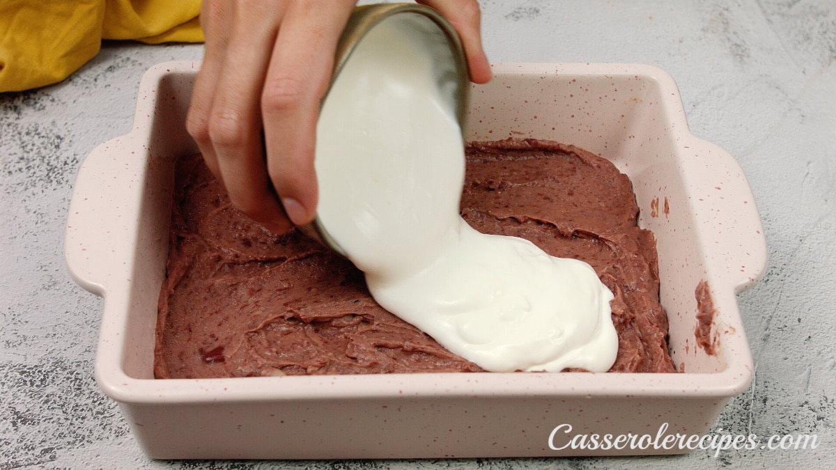 pouring sour cream over the refried beans in a casserole dish