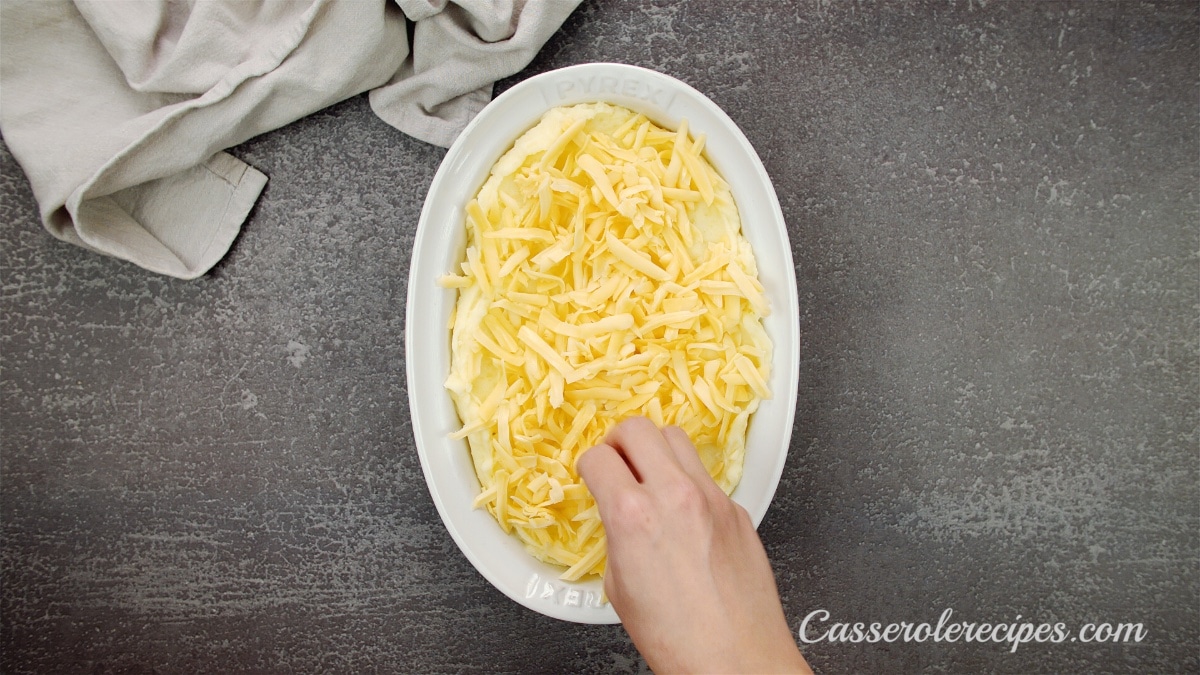 sprinkling cheese over potato casserole in a white baking dish