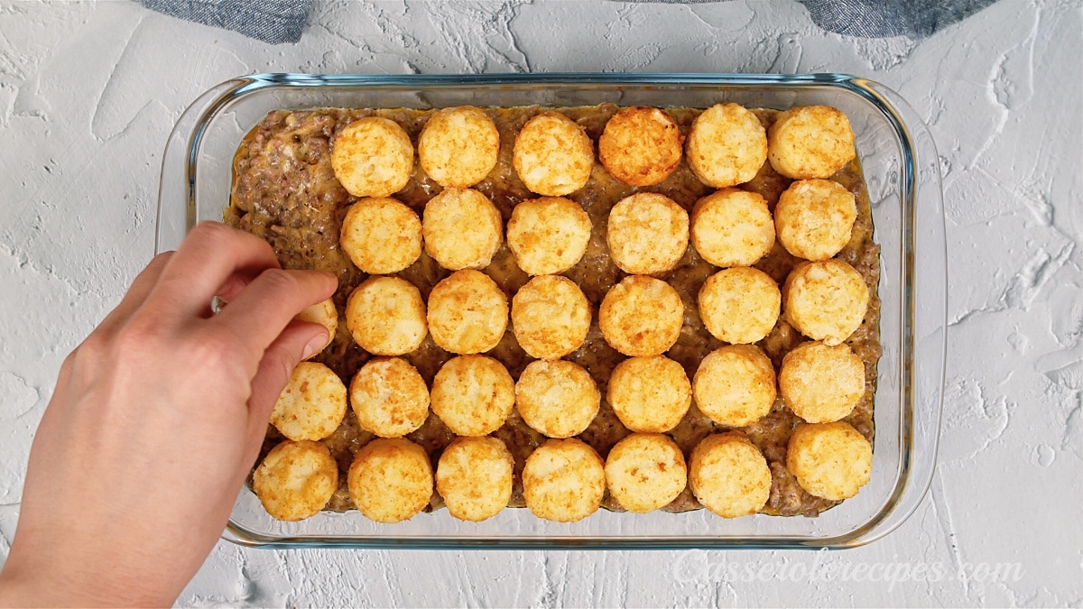 ground beef mixture in a baking dish being topped with tater tots by a hand