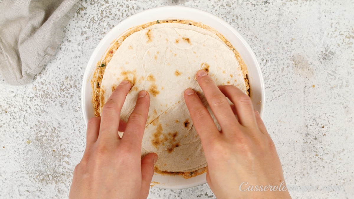 two hands holding tortilla in place over casserole