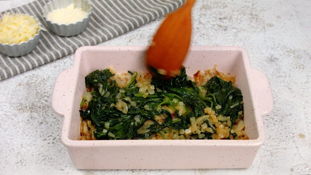 cooked spinach being spread over hashbrowns in baking dish