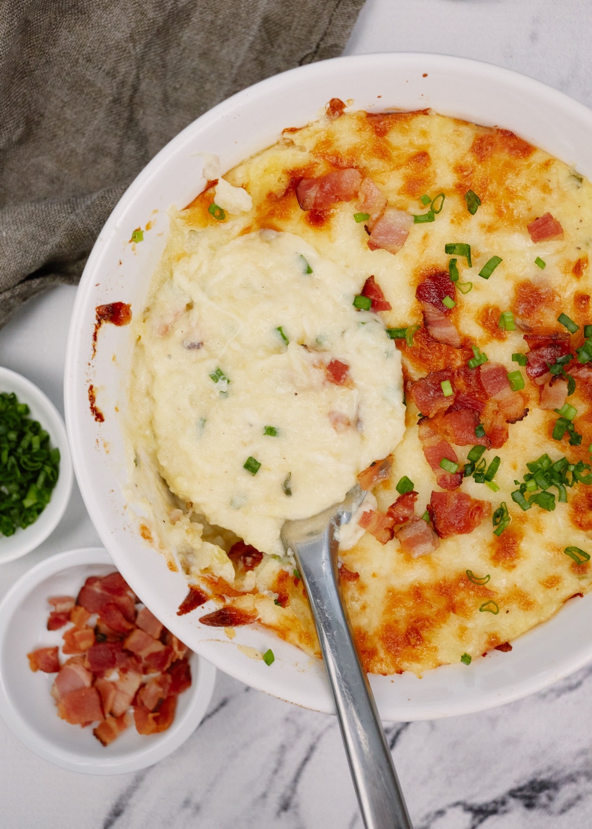 twice baked potato casserole in a baking dish with a spoon topped with bacon and chives