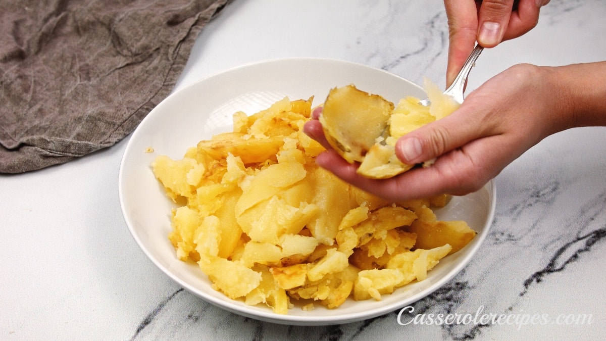 potatoes in a bowl with a hand using a spoon to scrape out potatoes from skin