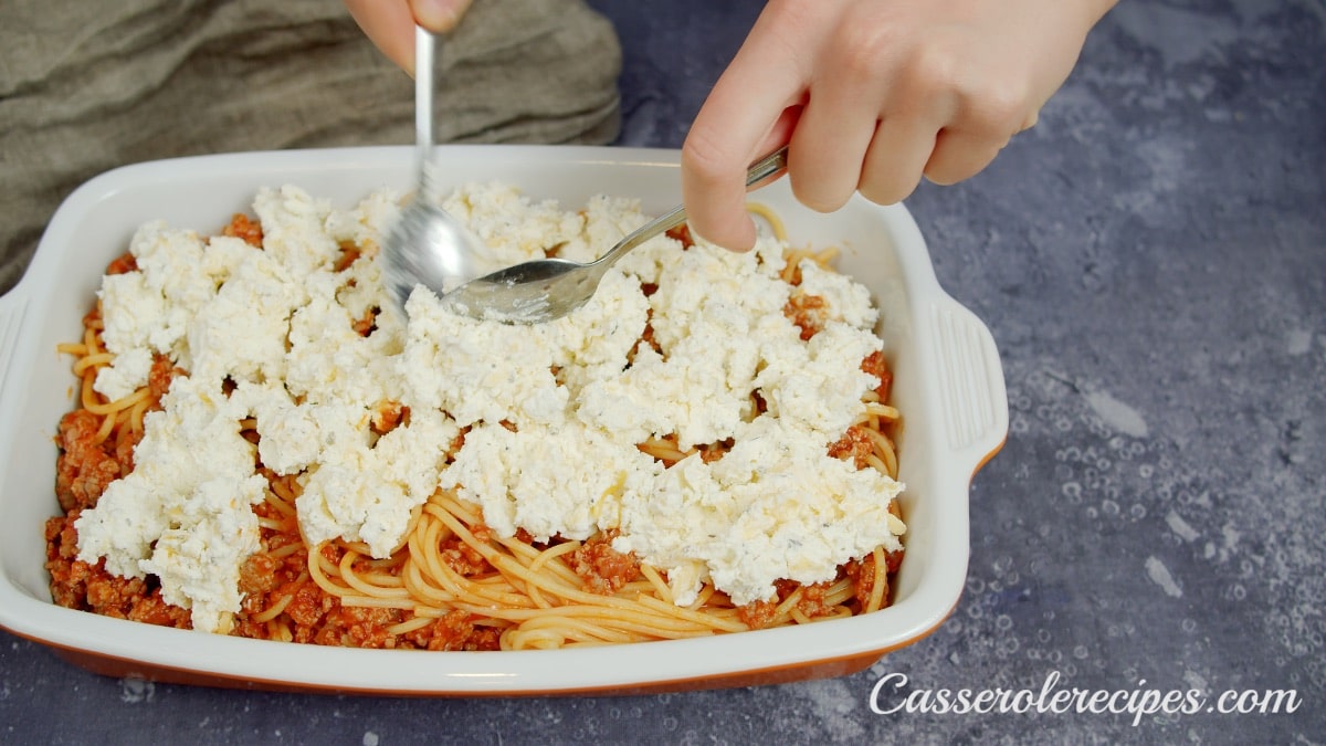 scooping small drops of cheese over spaghetti in baking dish