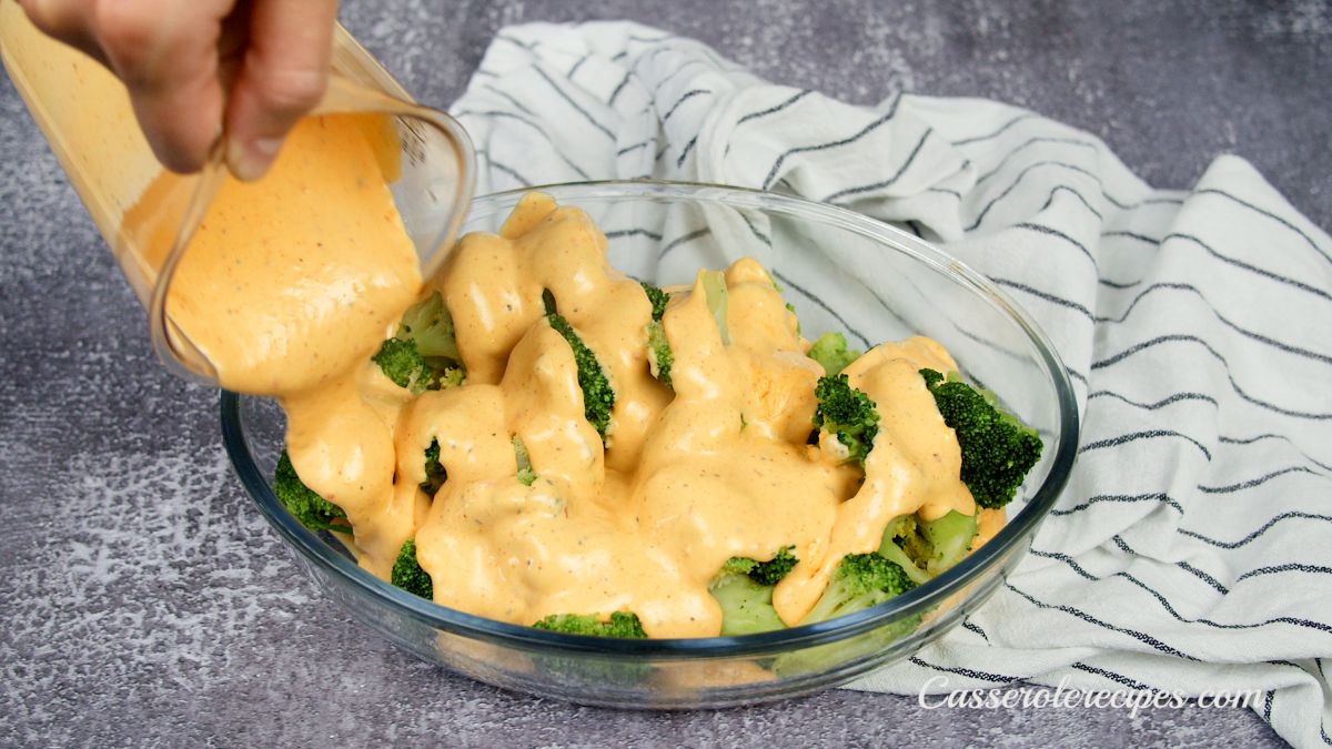 cheese sauce being poured over broccoli in glass baking dish