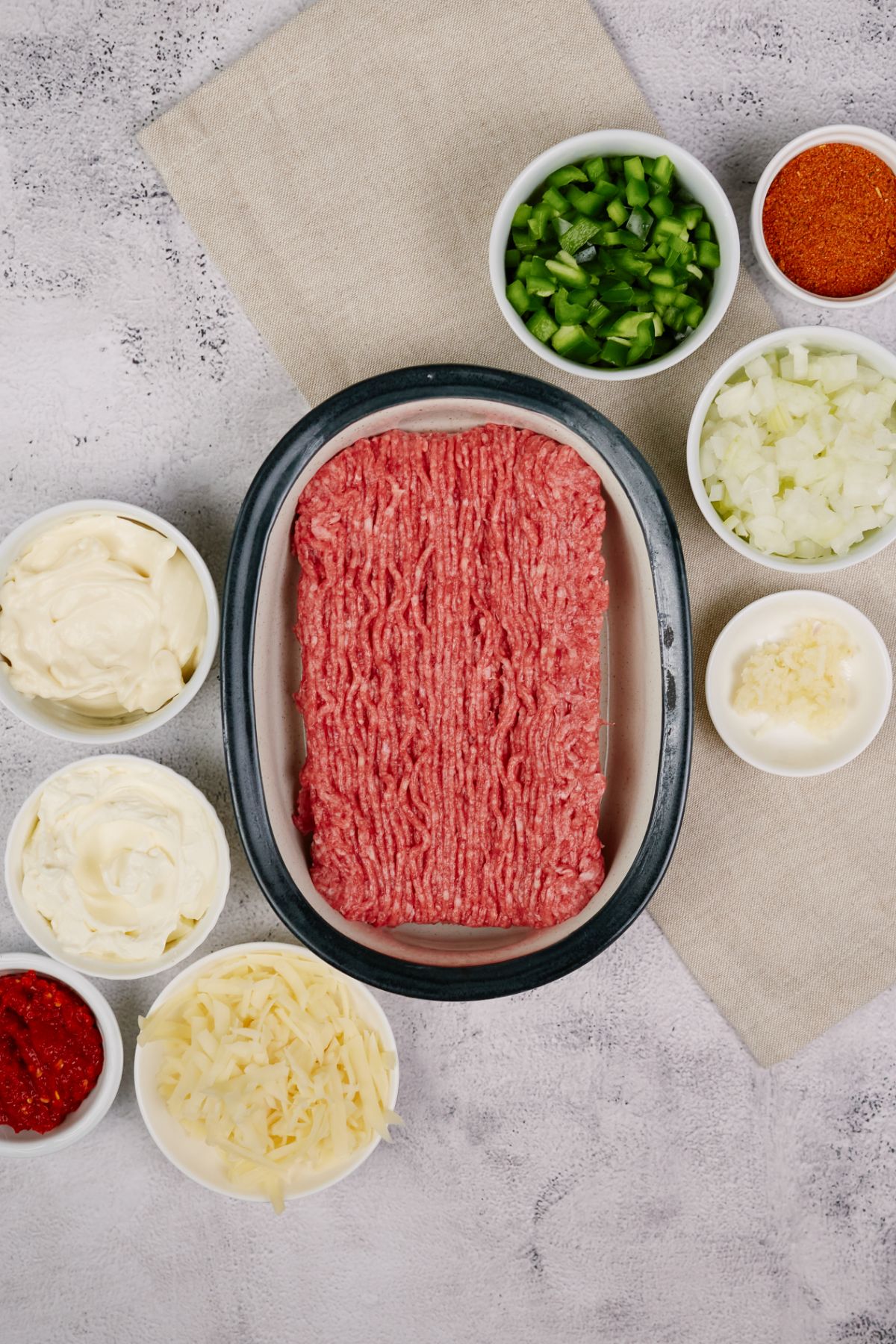 raw ground beef in large bowl next to bowls of cheese and vegetables on gray table