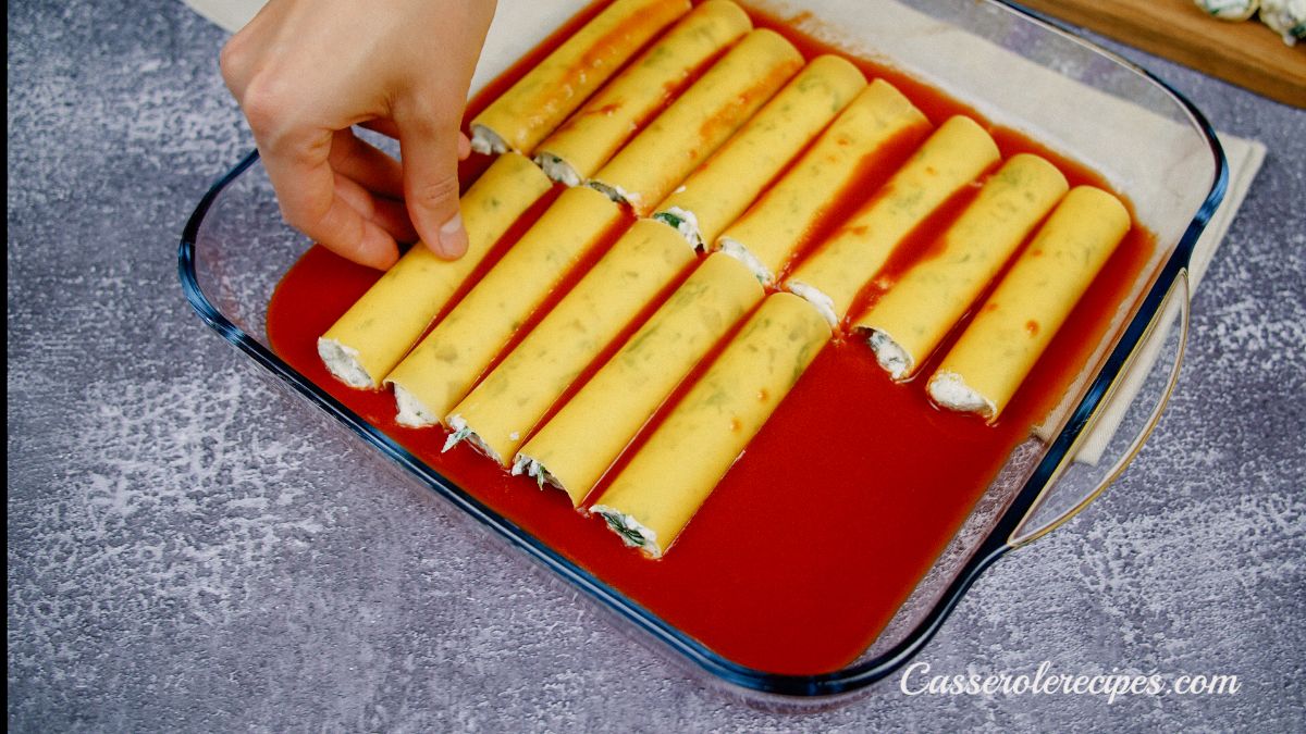 hand placing manicotti into baking dish with tomato sauce