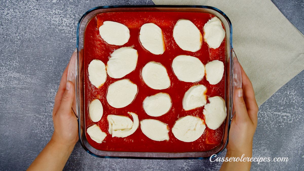 hands holding square casserole dish of manicotti topped with cheese