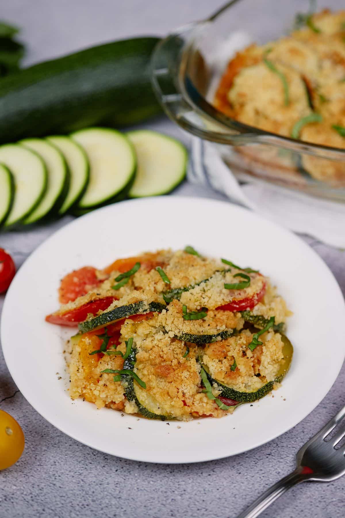glass casserole dish behind white bowl of zucchini and tomato with breadcrumbs