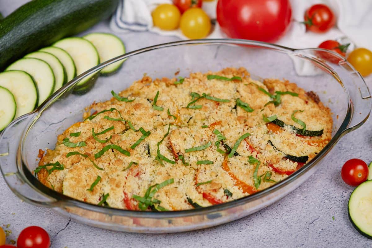 zucchini and tomato in casserole dish topped with breadcrumbs and herbs on gray table with zucchini and tomatoes in background