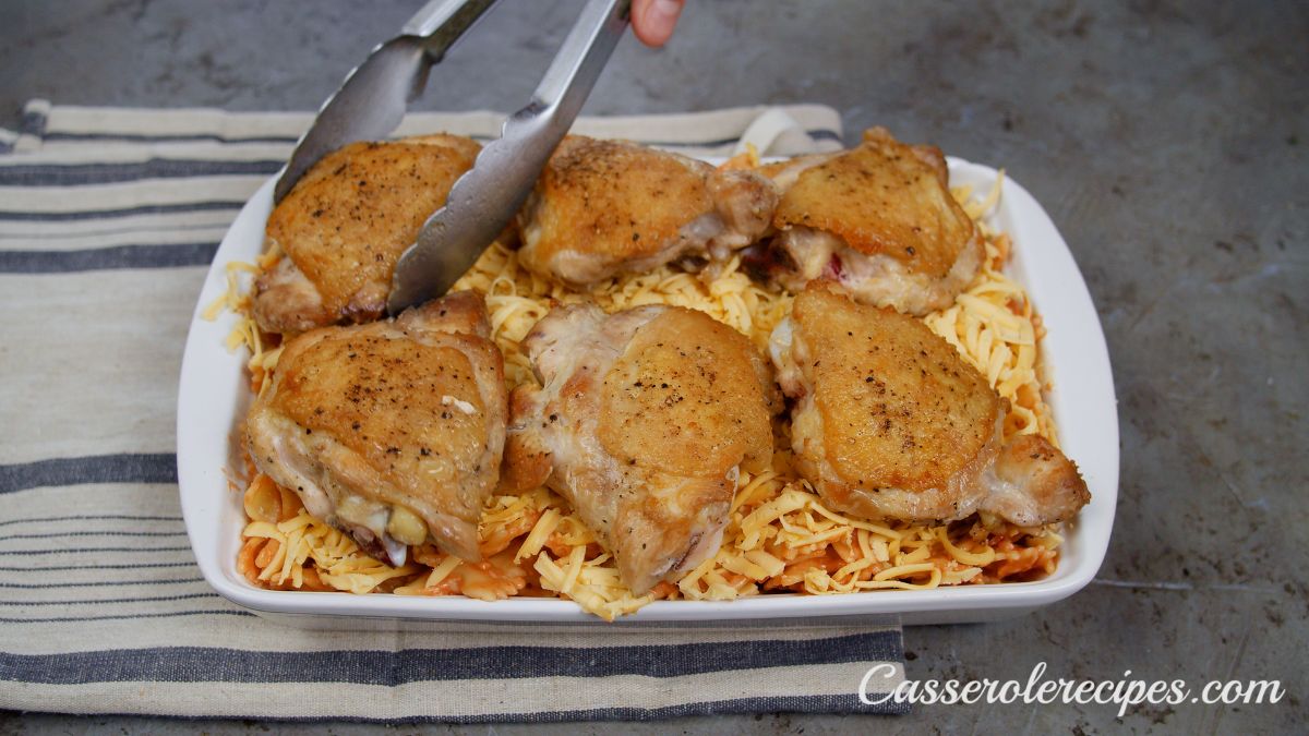 tongs placing par-cooked chicken thighs on top of pasta in baking dish