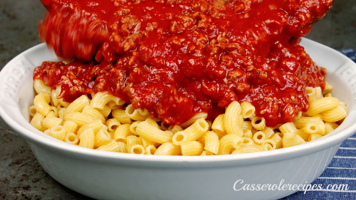red meat sauce being poured over cooked elbow macaroni in white baking dish