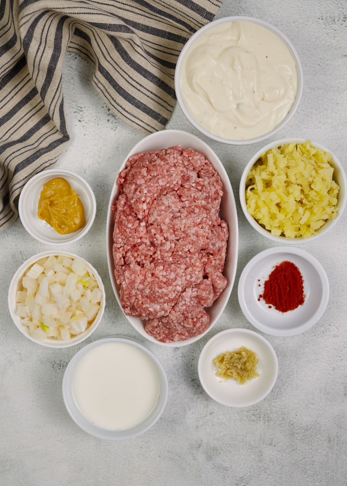 white ramekins and bowls of ingredients including raw ground beef on white table by striped napkin