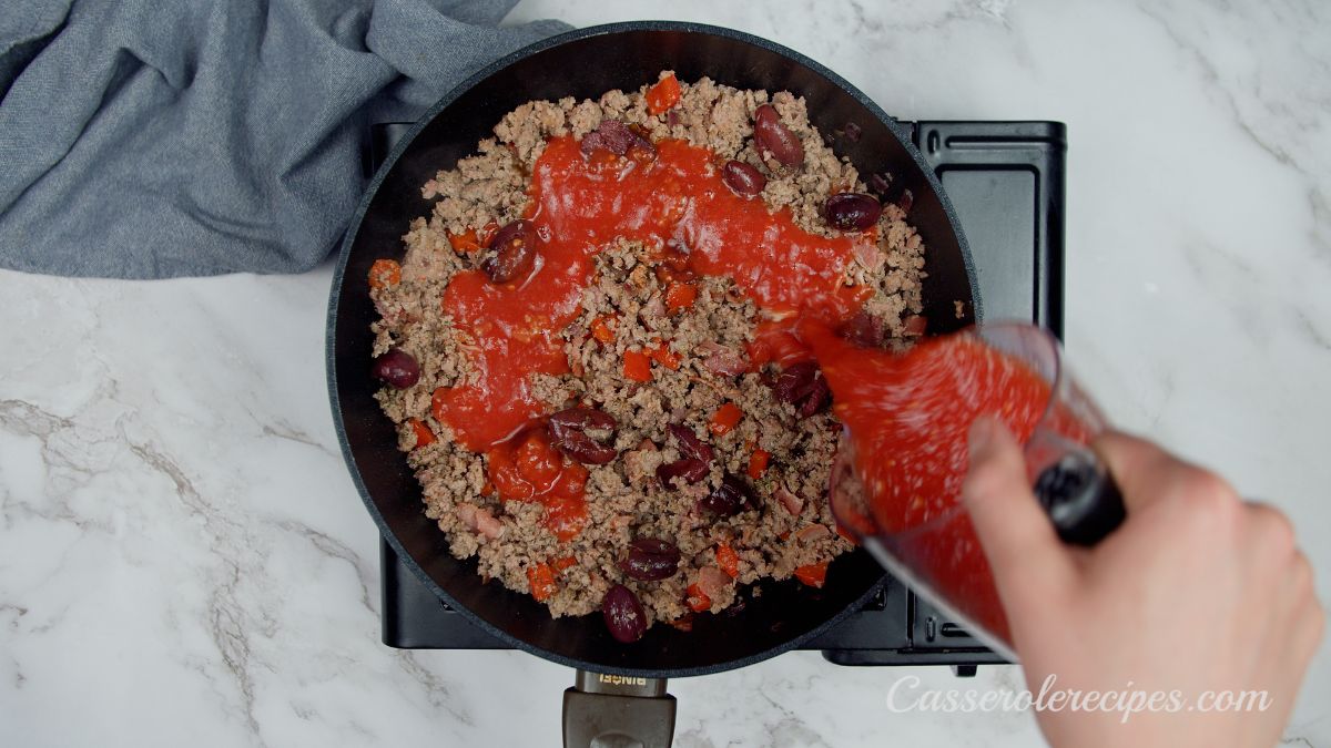 tomato sauce being poured into skillet with cooked ground beef