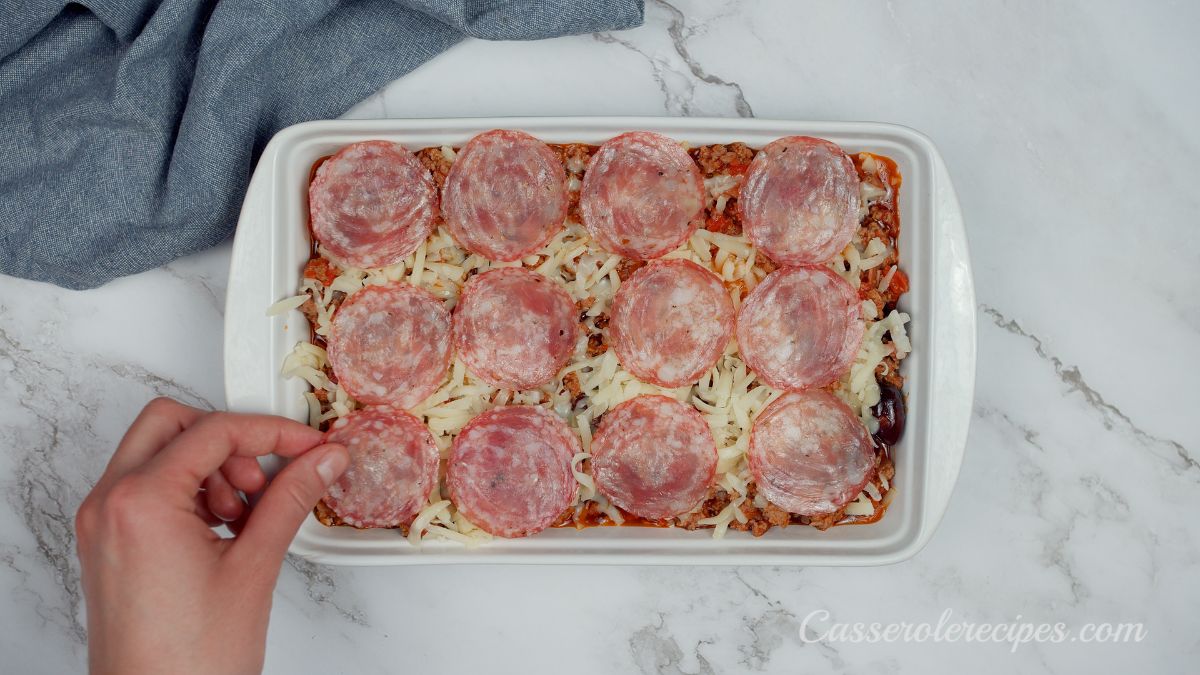 hand placing salami on top of cheesy casserole