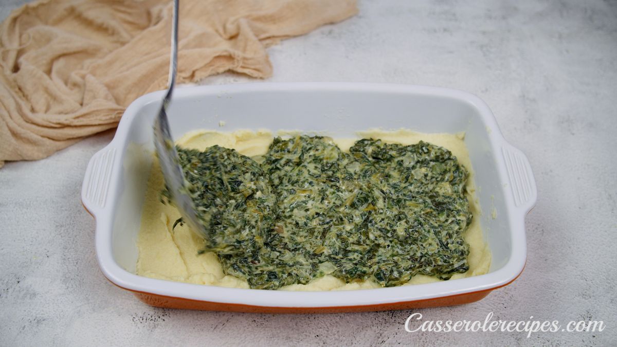 creamed spinach being poured over mashed potatoes in baking dish