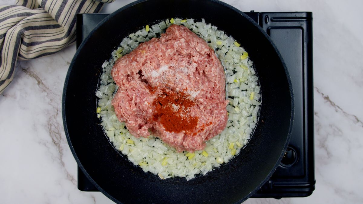 raw ground meat and spices on top of cooked onions in skillet on black hot plate