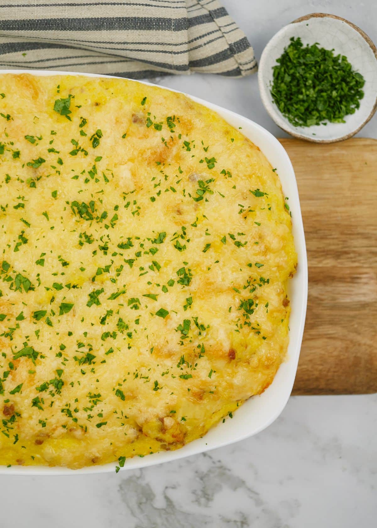 white baking dish filled with cheesy casserole on cutting board net to bowl of herbs