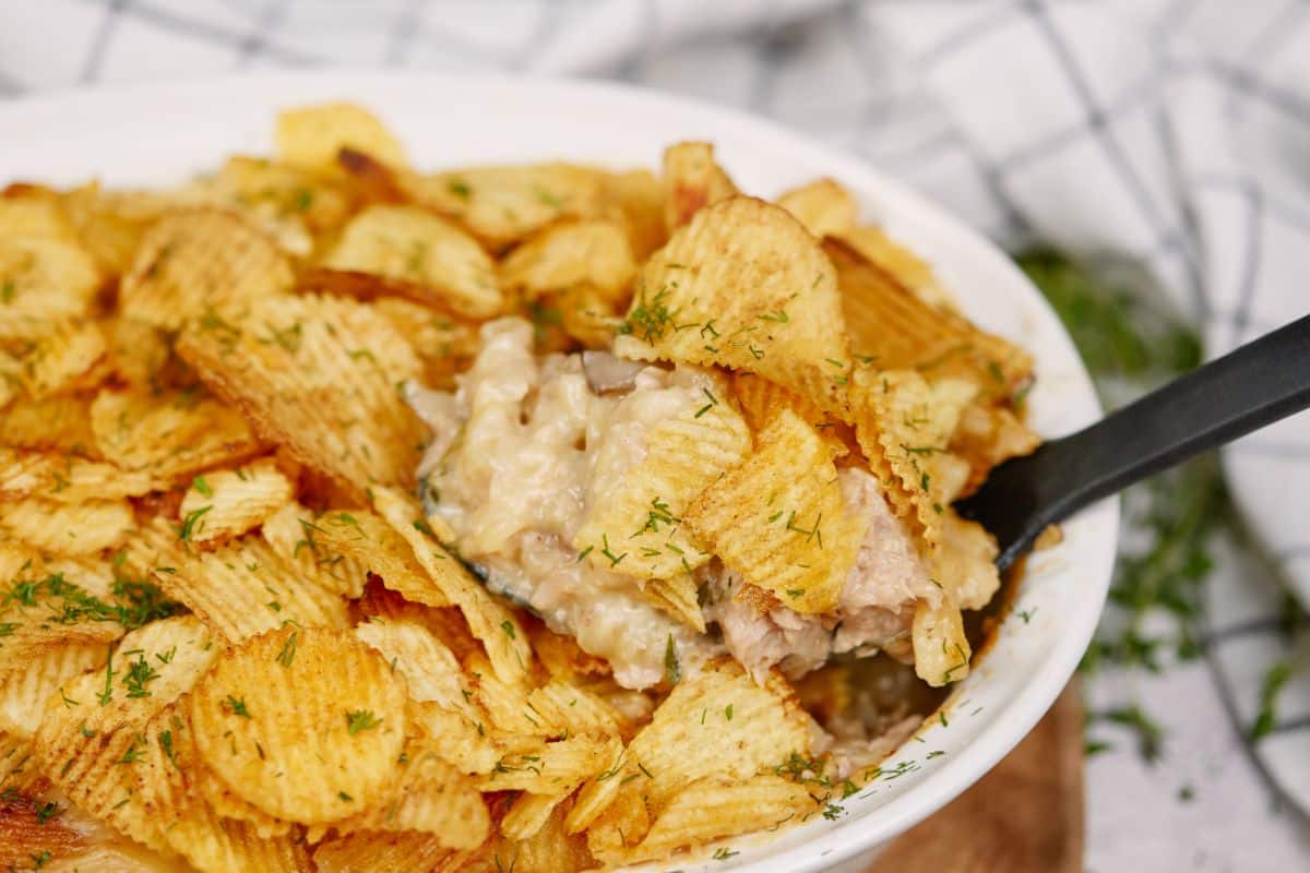 white casserole dish of tuna casserole with potato chips and dill on top and black spoon in side of dish