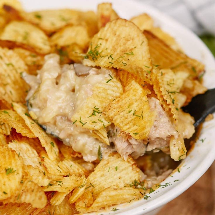 white casserole dish of tuna casserole with potato chips and dill on top and black spoon in side of dish
