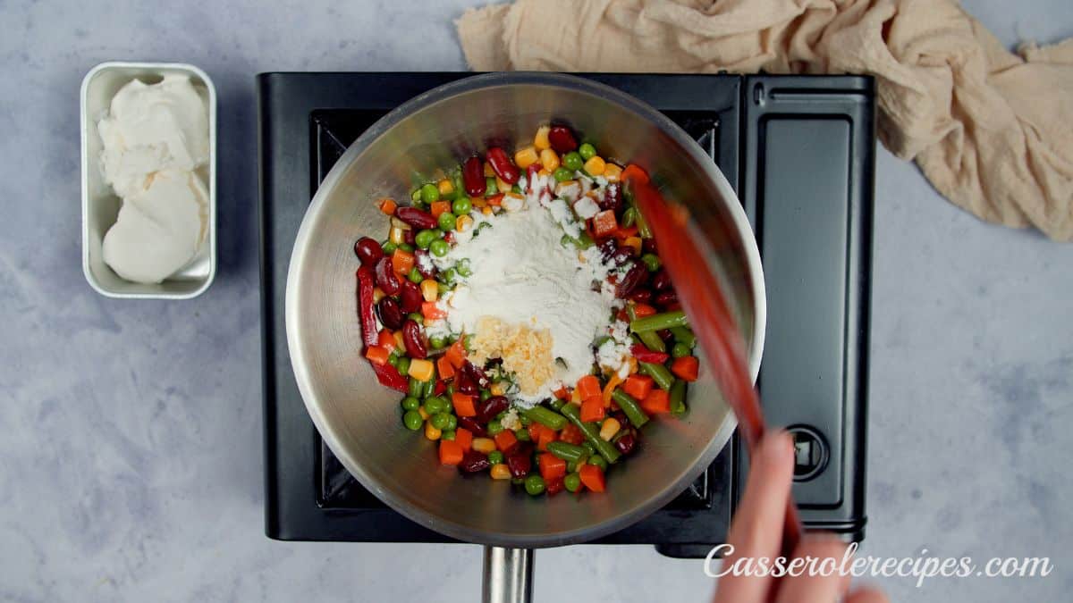 flour on top of mixed vegetables in skillet being stirred by wooden spoon