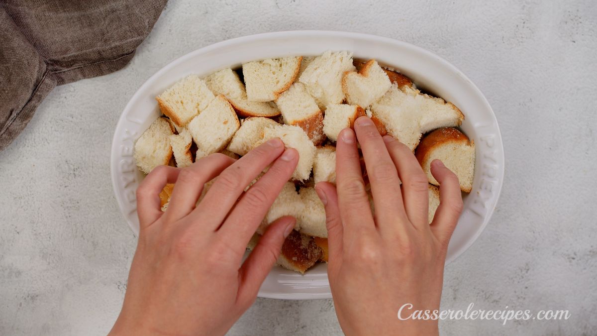 hand putting bread cubes into white oval baking dish