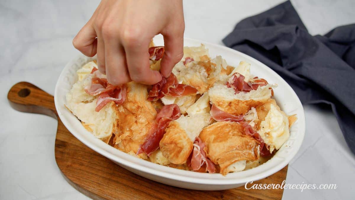 ham being put on top of croissants in baking dish