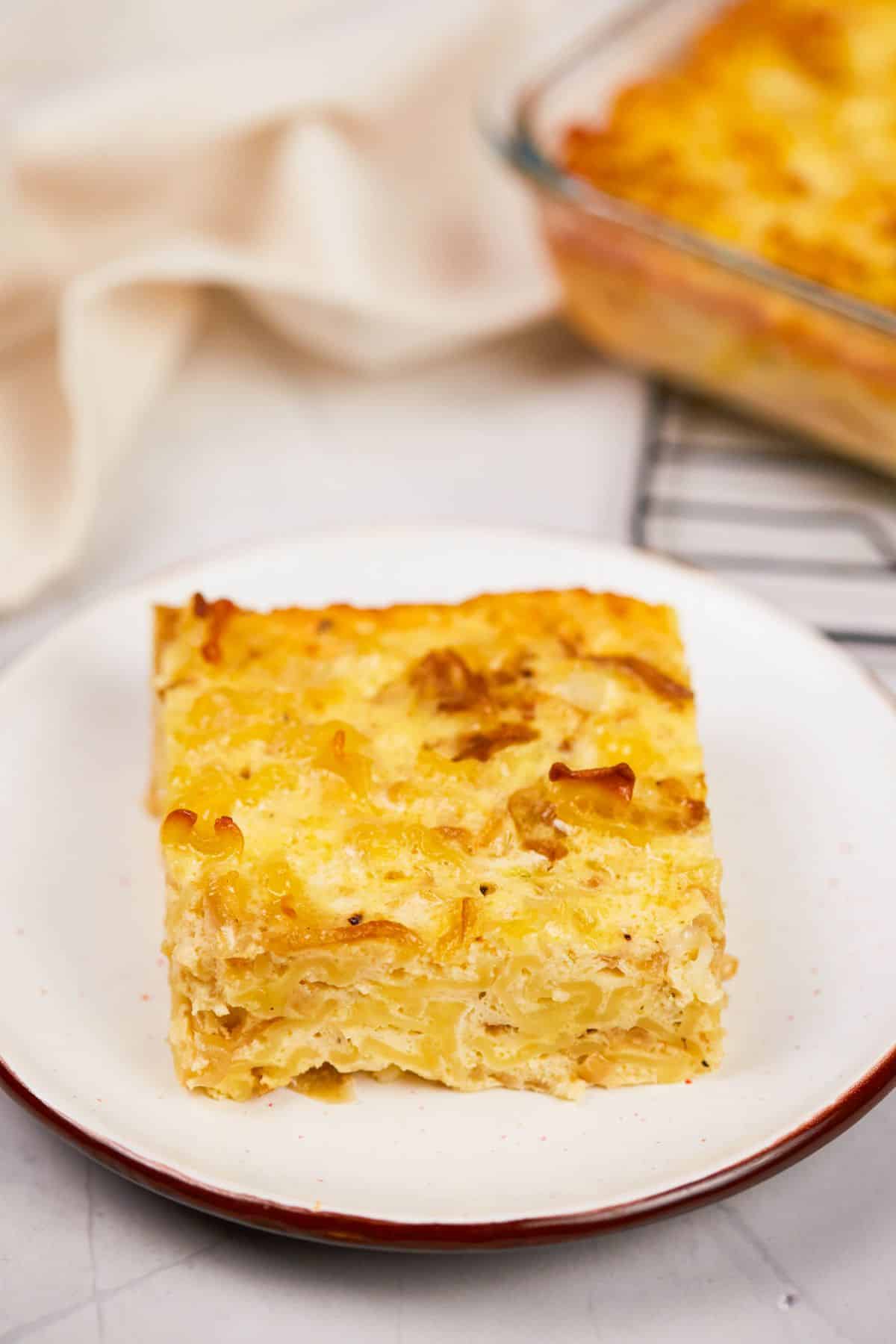 square slice of noodle kugel bake on round white plate by glass baking dish