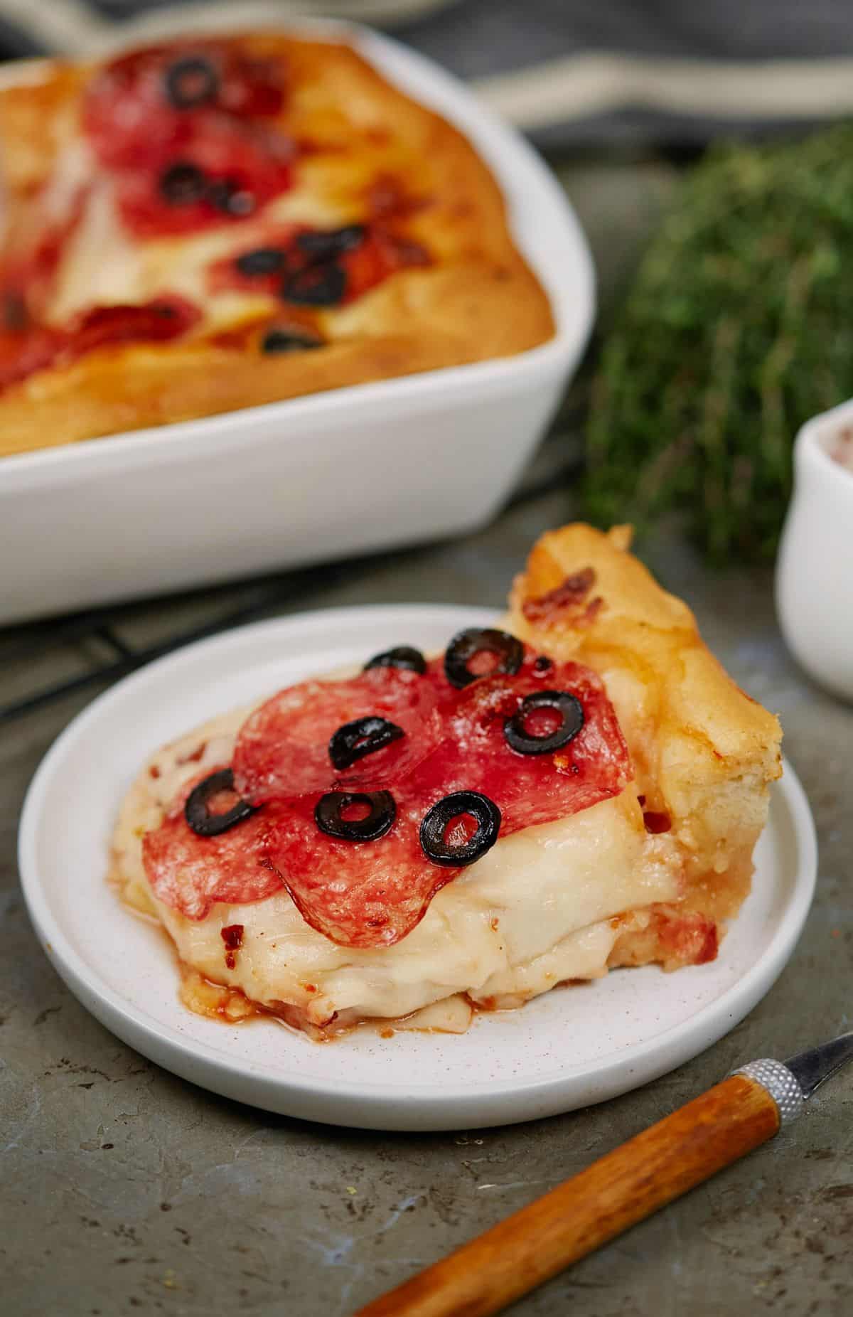 slice of deep dish pizza with salami and olive top on round saucer by white casserole dish