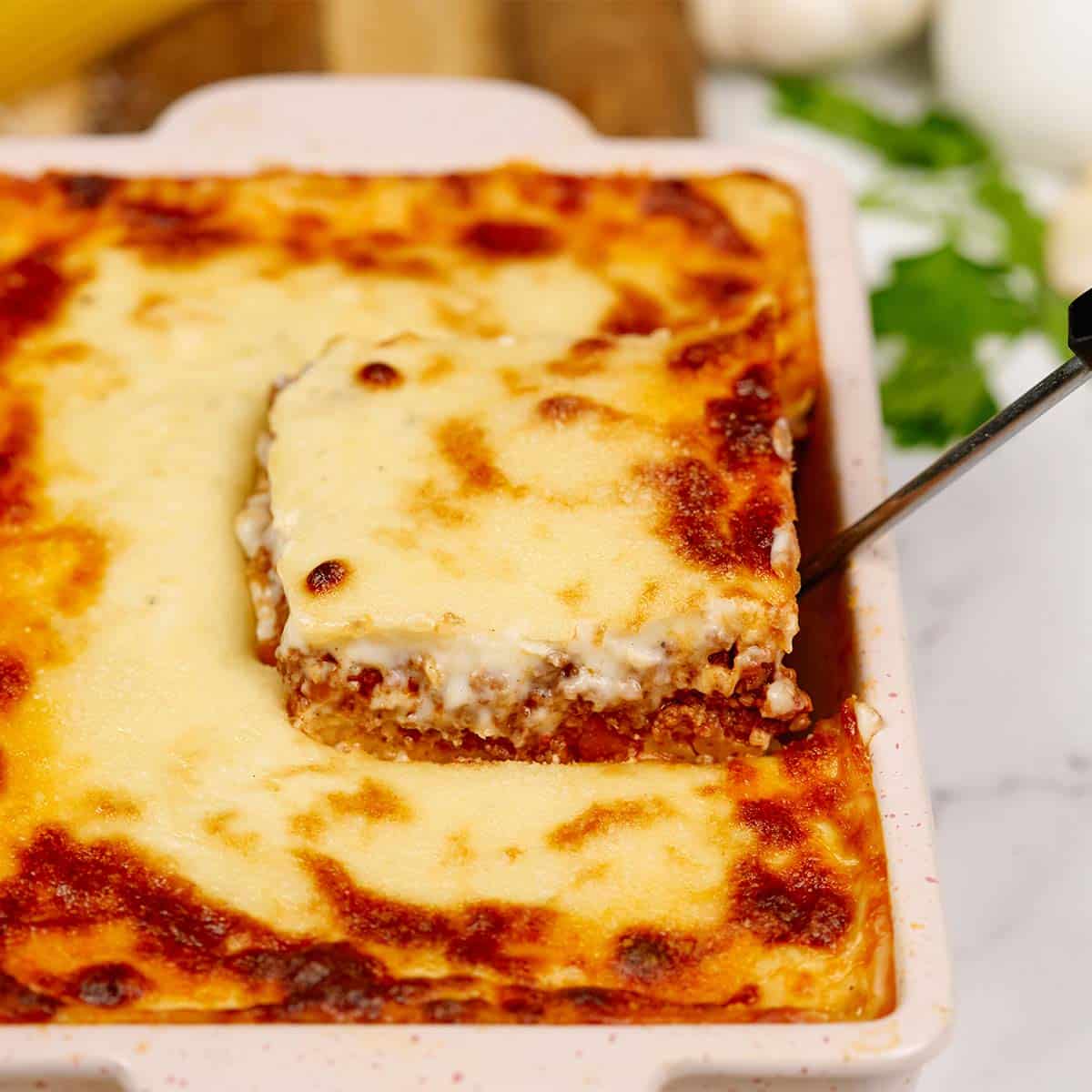 serving of pastitsio greek beef pasta bake being lifted from a casserole dish by a spatula