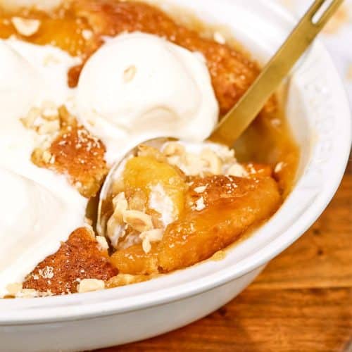spoon scooping up a serving of peach, almond and caramel self-saucing pudding from a casserole dish