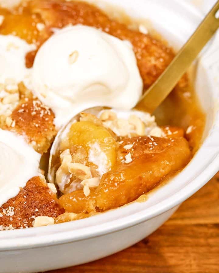 spoon scooping up a serving of peach, almond and caramel self-saucing pudding from a casserole dish